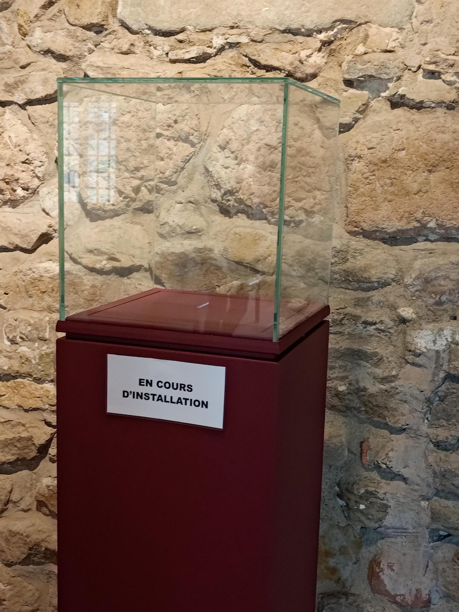 Un présentoir en bois surplombé d'une cube en verre devant un mur en pierre. Mais ce présentoir est parfaitement vide. Une écriteau sur le présentoir indique "En cours d'installation".